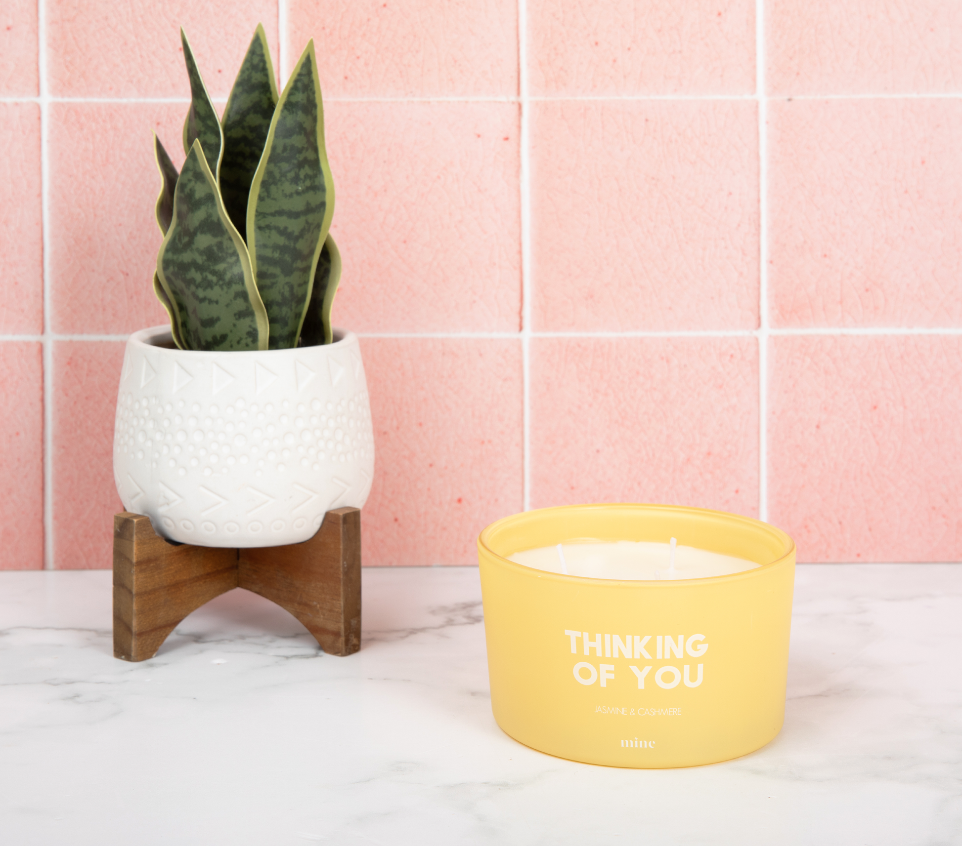 yellow candle glass that says Thinking of You placed on white marble top and pink tile background placed next to house plant